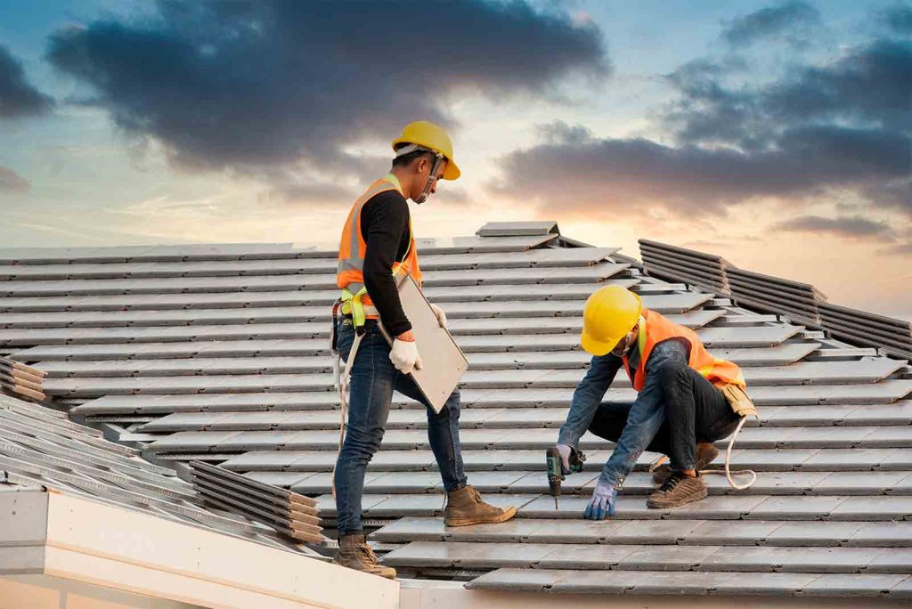 two construction workers on a metal roof: one stands with a clipboard, the other drills while kneeling, both in hard hats and reflective vests at sunset
